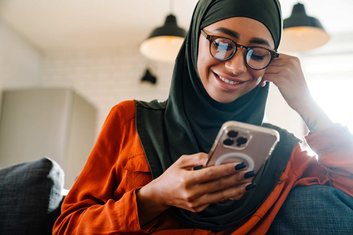 A smiling woman in glasses and a green hijab looks at her smartphone in a cozy, modern home