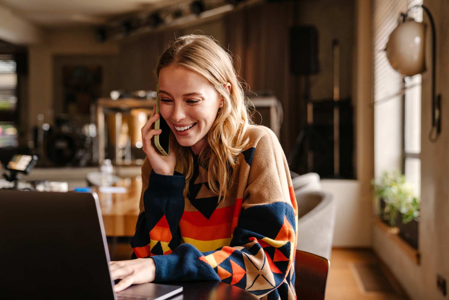 A smiling woman in a colorful sweater talks on the phone while using a laptop in a cozy, modern room
