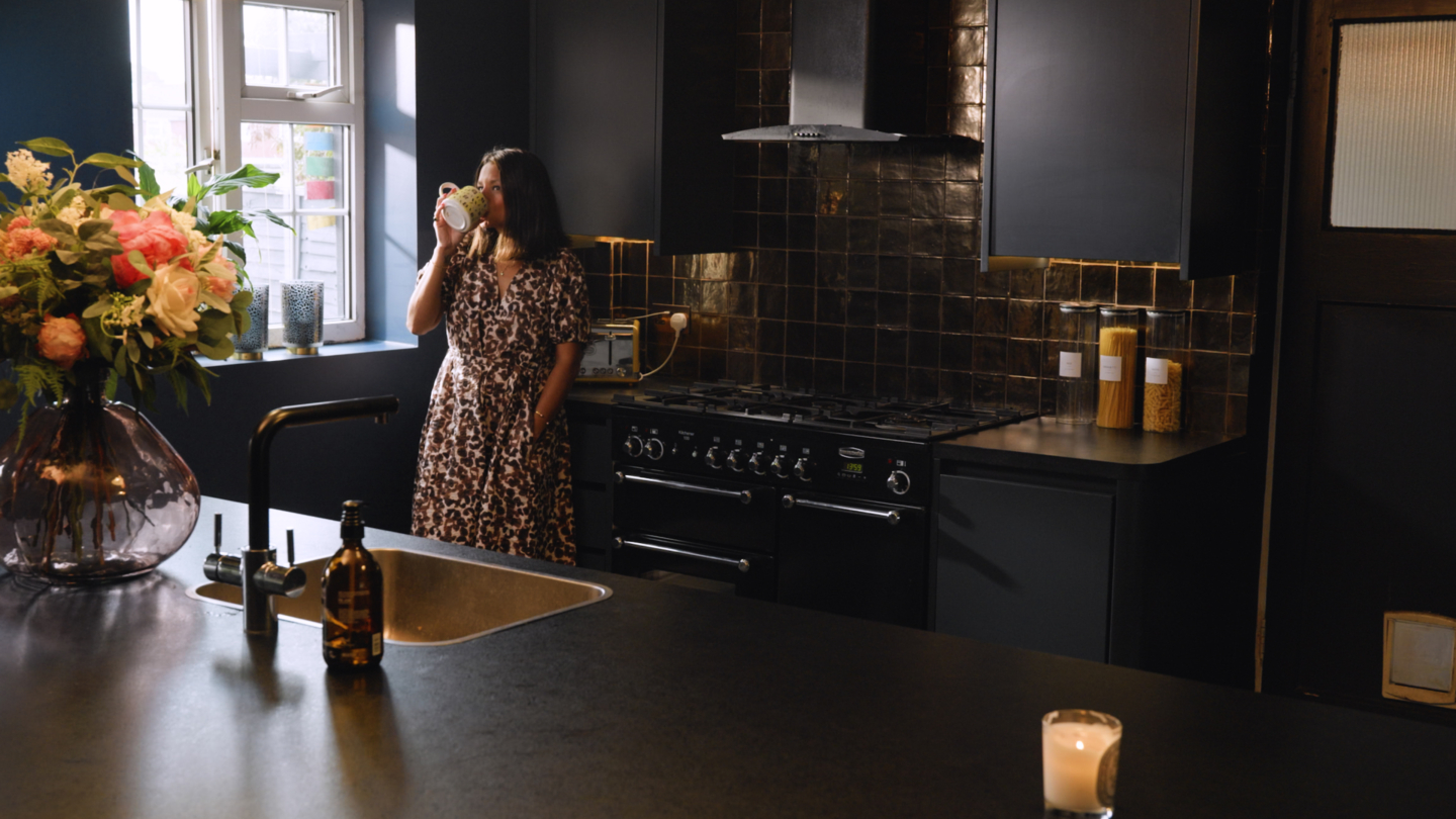 A person drinking coffee in a kitchen, with black worktops, black tiles and a range cooker
